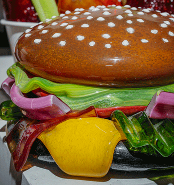 Life-size glass hamburger with bun, overflowing with green lettuce, red onion, green pickle, bacon slide and yellow cheese.